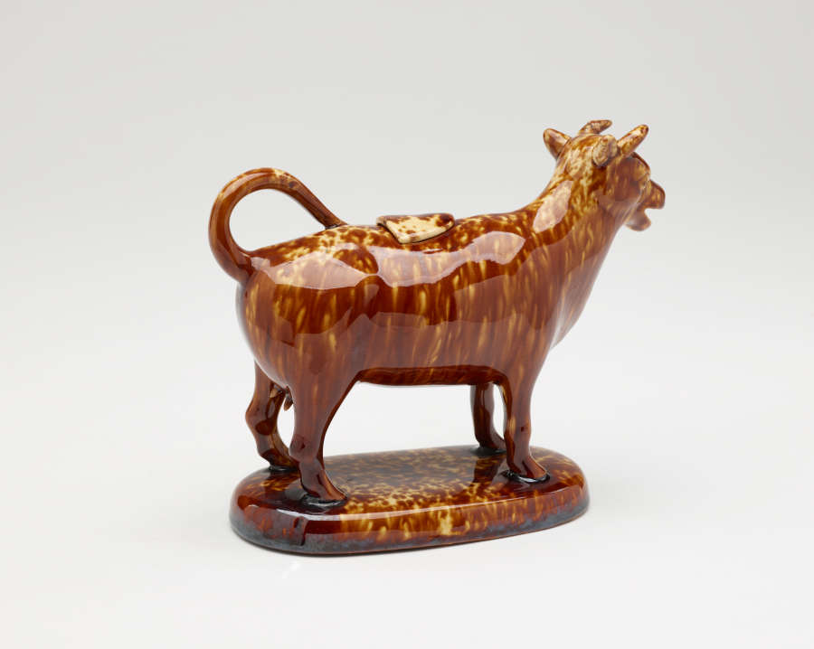  A brown sculptural creamer in the shape of a cow with horns. There is a small flat lid in the center of its back.