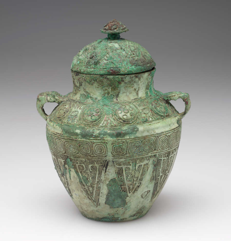 Opposite side view of a vessel with a round bulbous body, short neck, round lid, and two handles. The surface features patches of different greens, and carvings of geometric patterns.
