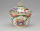  A white bowl with lid and finial with green, red, blue, pink, yellow and gilded decorations. The finial is a sculptural flower white, red and green.