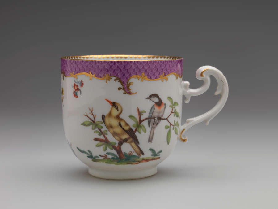 A white teacup with a swirling handle. Gold and pink decoration at top edge. Central design features two birds on a branch.