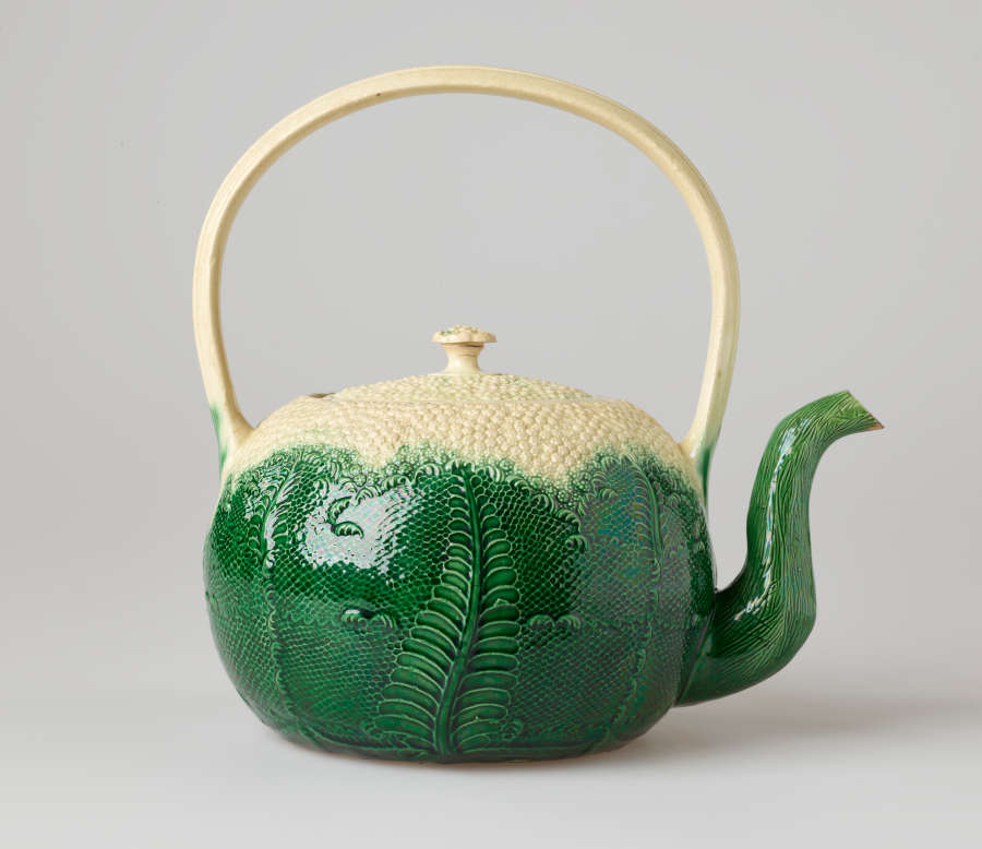 A large pot in the shape of a cauliflower with a green base and white top. A tall handle arches directly above the lid.
