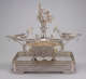 Profile view of elaborate silver double-oval centerpiece with swirling arms extending to two shallow side-dishes and central fountain-like stem topped by classical figurines, all on a rectangular tray pedestal.  