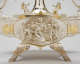 A silver oval relief panel depicting a central flowing-robed woman surrounded by playful cherubs and swirling drapery, enclosed in a gold-trimmed frame attached to a tray elevated by ornate legs. 