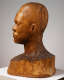 Profile view of a wooden bust depicting the head of a male figure with afrocentric facial features. His expression is stern and holds an intense gaze. The wood is cracked.