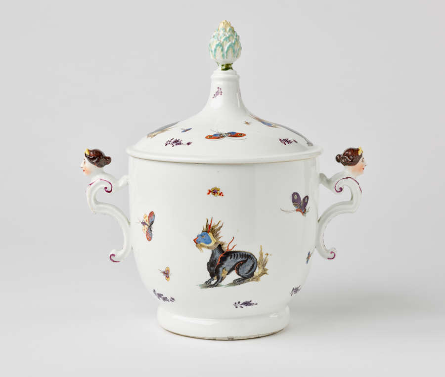 A white lidded vessel with dragons and bugs decorations. There are two sculptural handles with figure’s heads at the top. Finial has light green, leaf-like decorations.