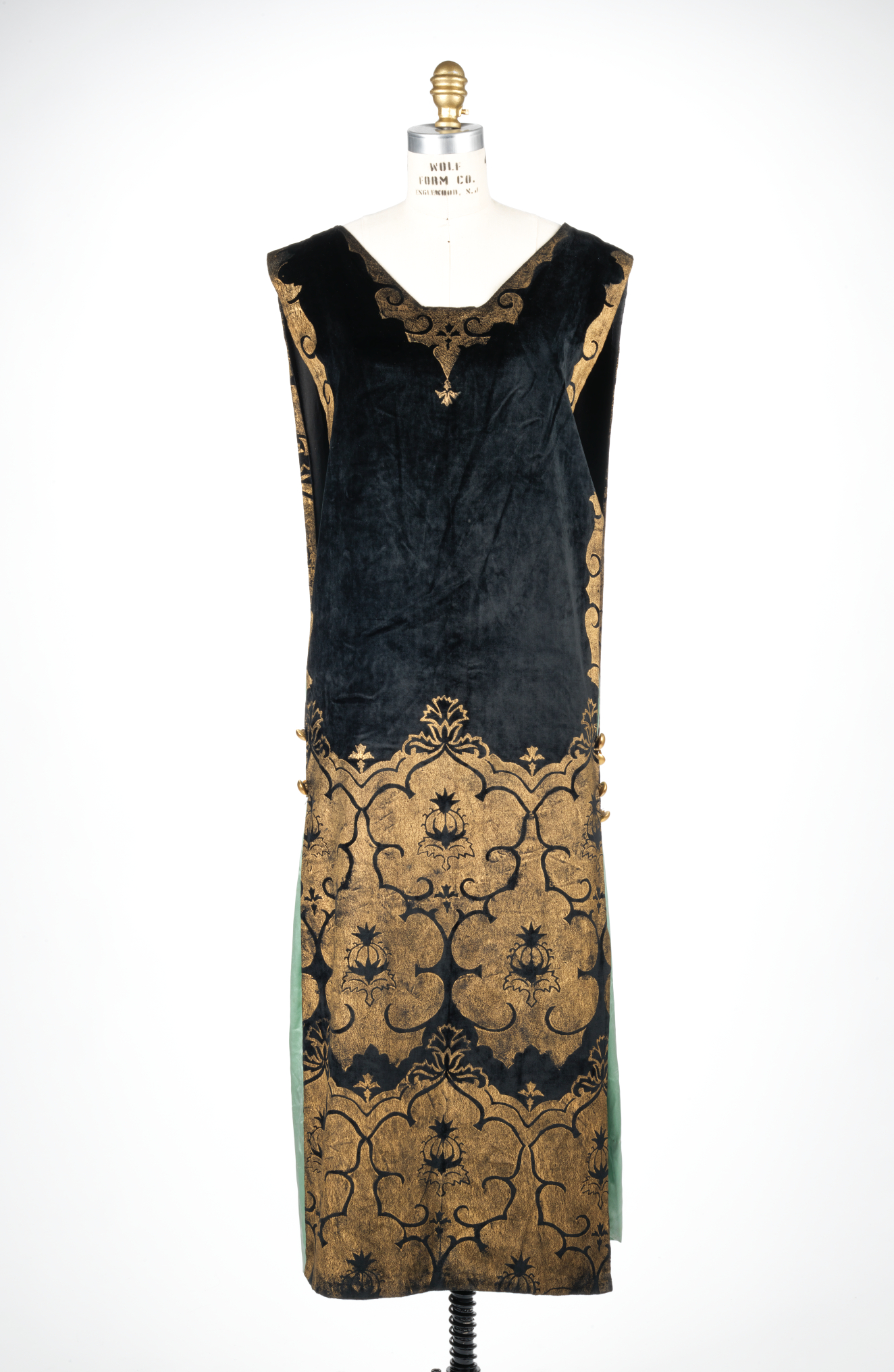 Front view of a sleeveless V-neck rectangular black dress with bold metallic gold floral patterning on the bottom half and shoulder hems, with pale green peeking from the side slits.