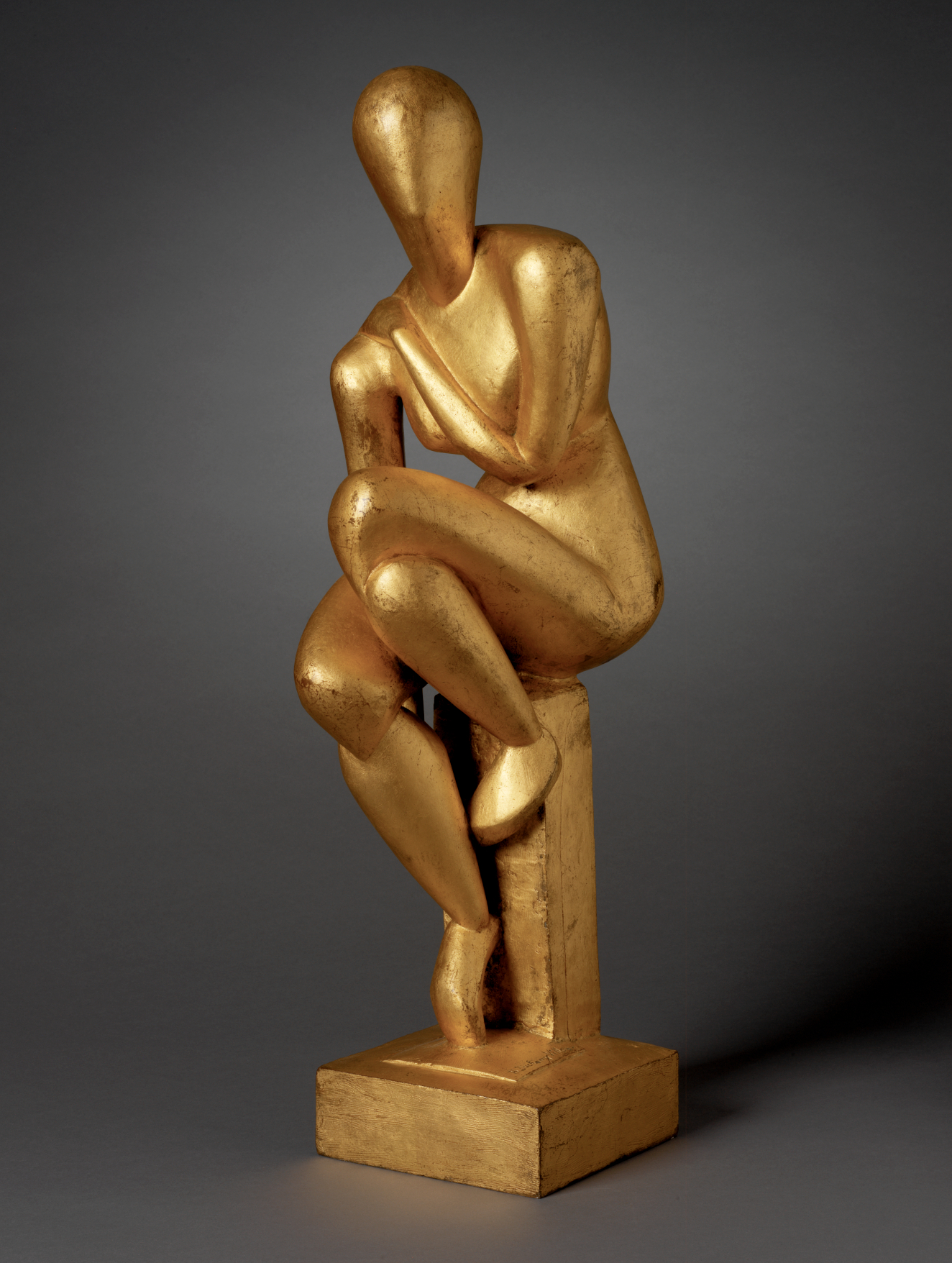 A golden sculpture of a faceless figure seated on a pedestal. Its smooth, elegant limbs posed with one arm over its chest and the other resting near its raised knee. 