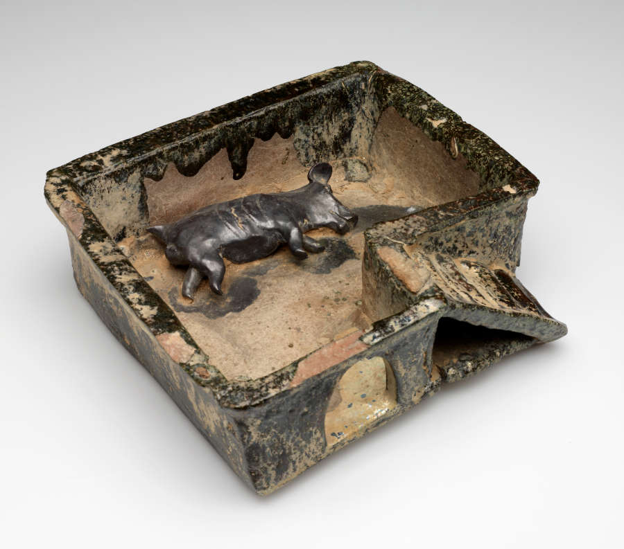 Ceramic sculpture showing a small pig laying on its side, enclosed by a rectangular structure which features arches and stairs. The sculpture is tan with a glossy muted green glaze.