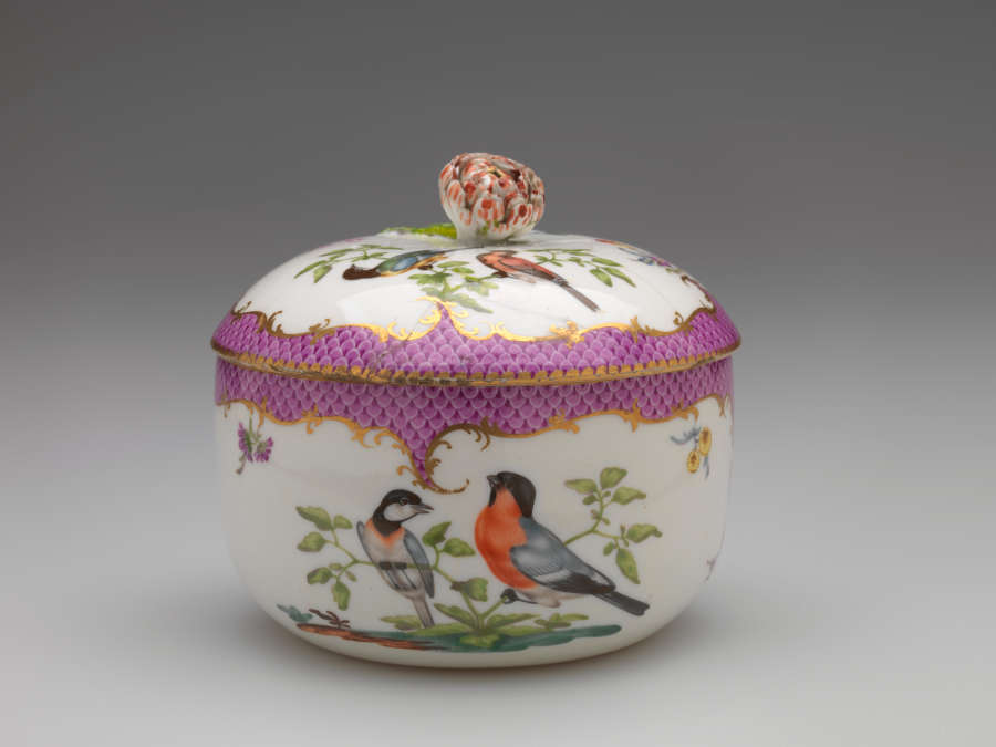 A white vessel with a lid, both with gold and pink edged decoration. Lid and body of vessel are decorated by two birds sitting on a branch.