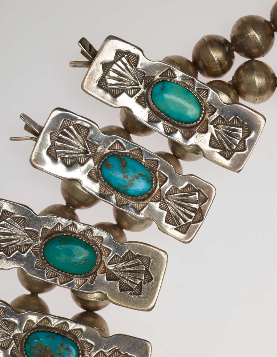 Detail-view of ornaments threaded into the necklace’s beaded chain. The rectangular ornaments have turquoise stones set in their center with silver decorative engraved borders on either side.