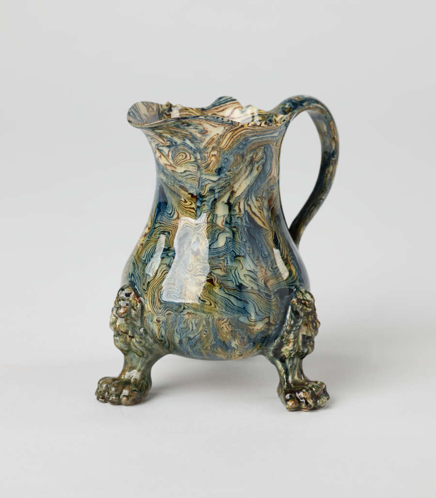 A small vessel with a cream, blue, and green mottled design. Three sculptural feet are shaped like lion’s paws with lion heads attached to body of vessel.