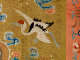 Embroidered textile featuring a crane with a red crown and outstretched black and white wings soaring among blue and pink clouds, set against a golden background.