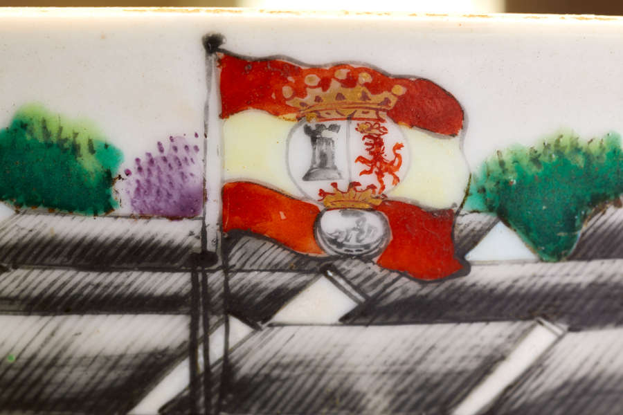 A close up of a ceramic of a red and yellow striped flag with a crown, a lion, and a castle emblem, against a background of black rooftops and foliage.