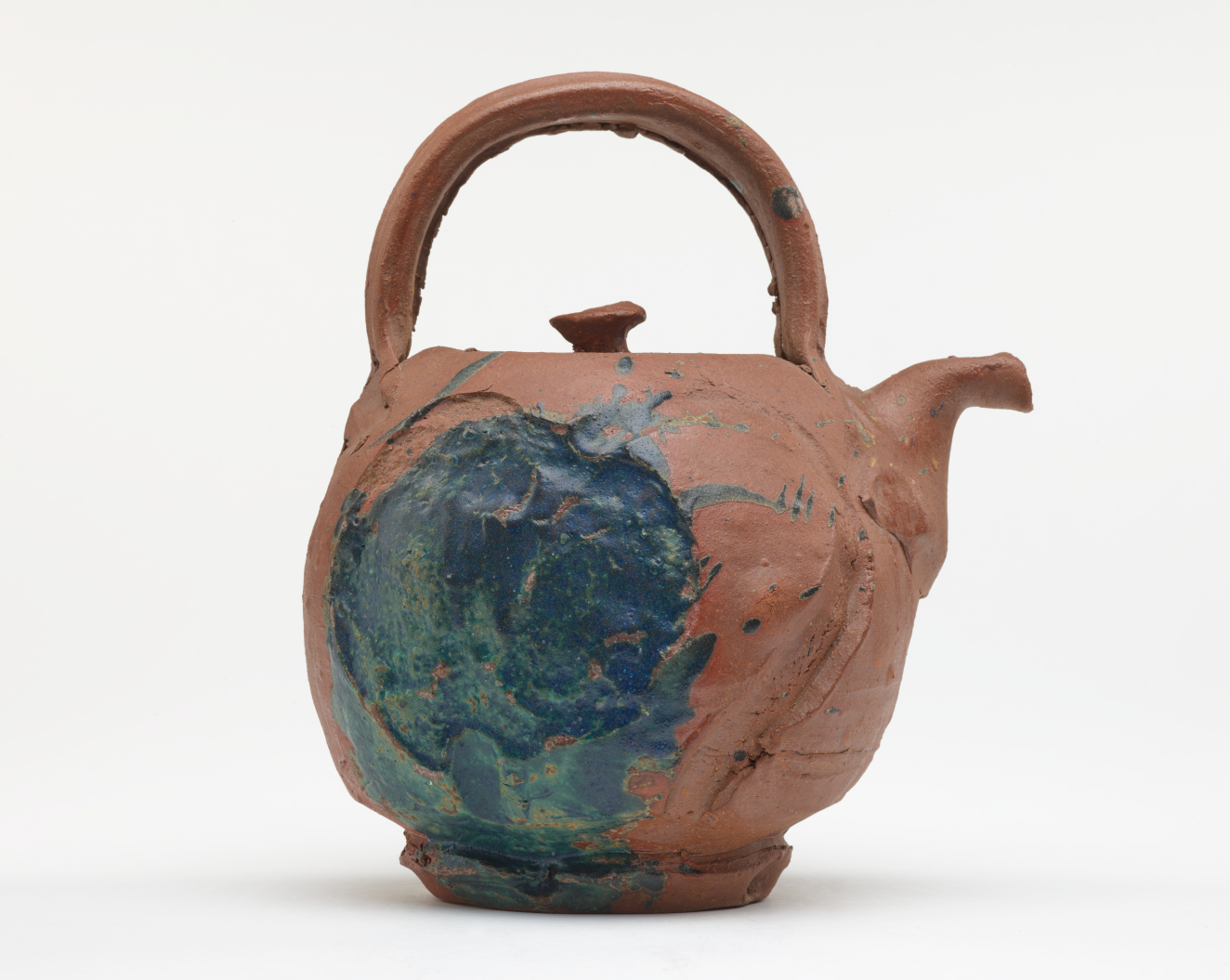 Brown clay teapot with a rough, uneven surface, a wide overarching handle, a short spout bent at a right angle, and a thick circular patch of blue-green glaze.