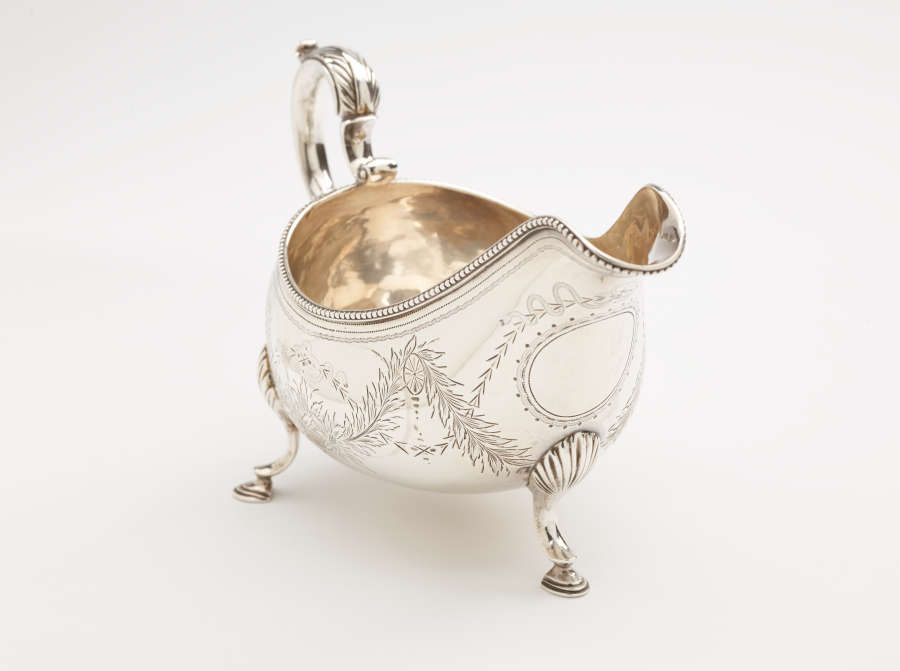 A silver sauce boat with a sculptural handle, high lip for pouring, and three protruding feet. There are floral engravings along the body of the vessel.