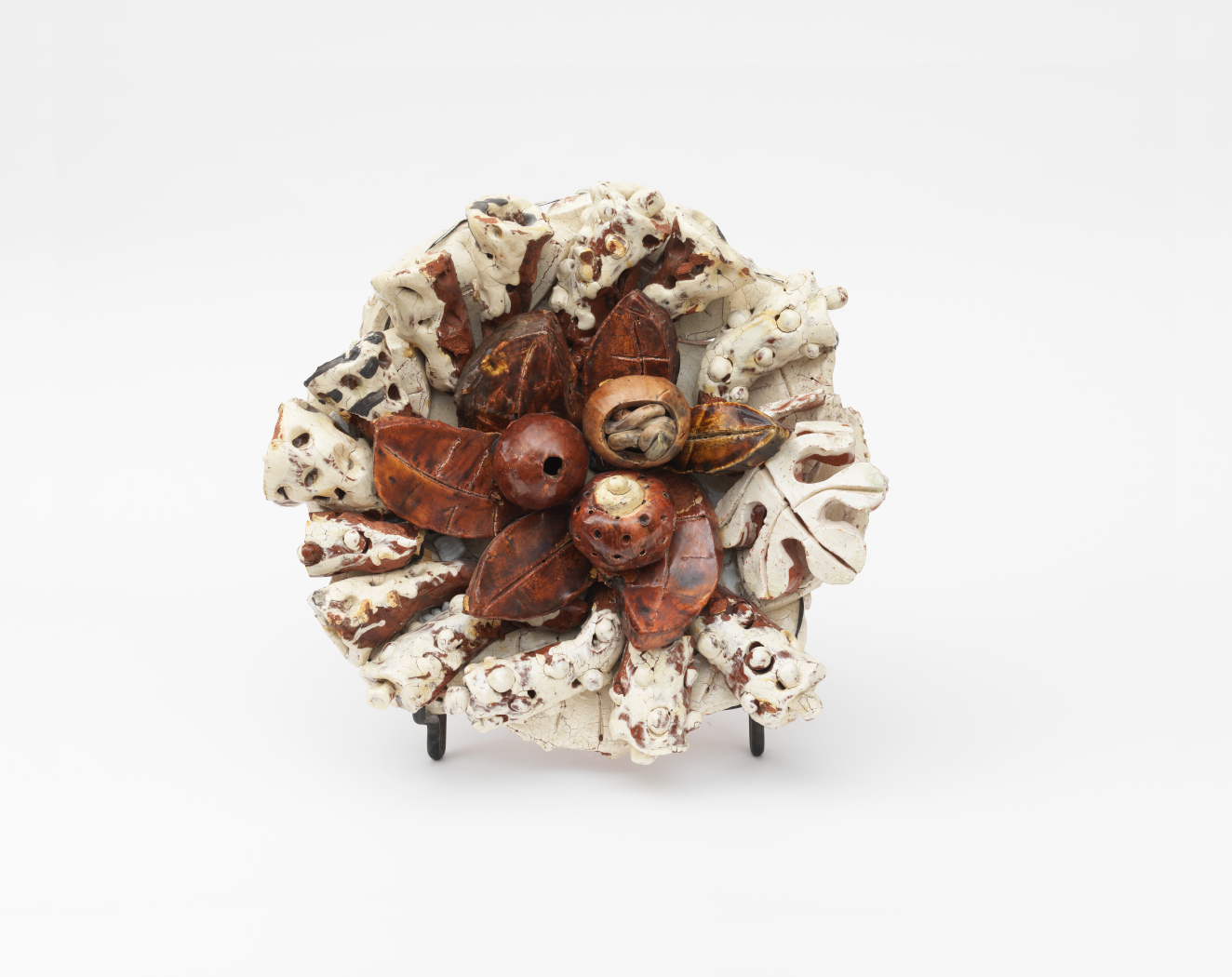 Circular ceramic sculpture featuring a cluster of glossy brown, seed-like pods at the center, surrounded by textured cream leaves speckled with brown radiating outwards from the cluster of seeds.