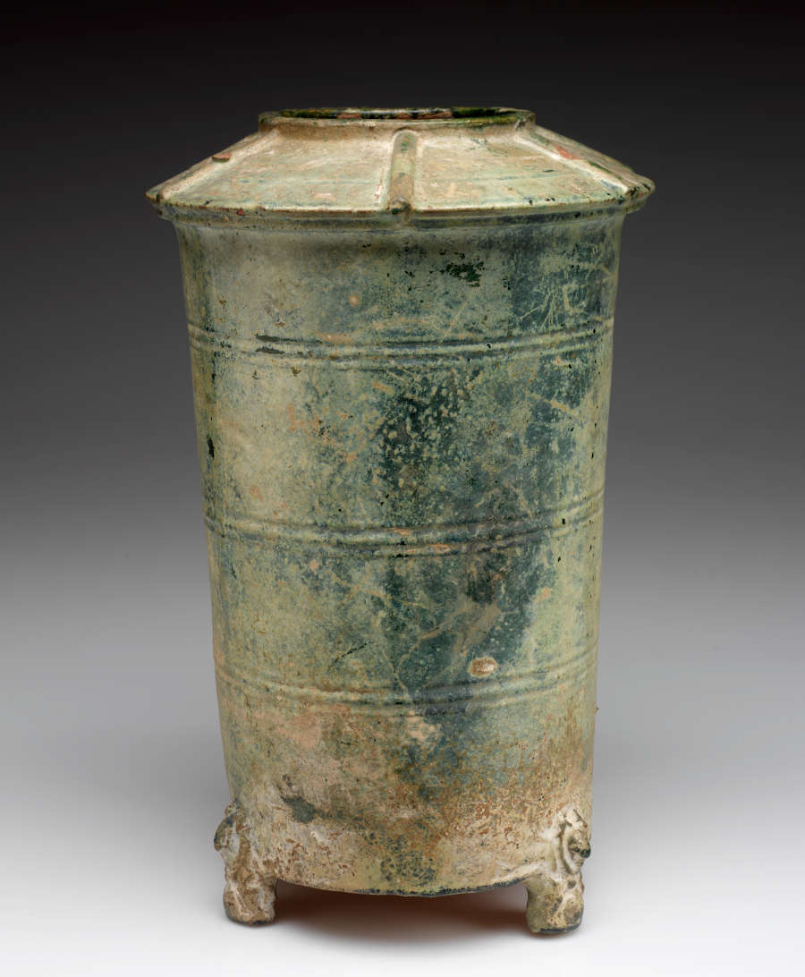 Side-view of a tall and narrow green-gray cylindrical terracotta urn with a narrow mouth and wide, upturned saucer-like neck, tapering into a narrow base, with one ornately carved foot visible.