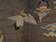 A close up of an embroidered white crane with outstretched wings, a red crown, and green legs, soaring against a gold lattice background.