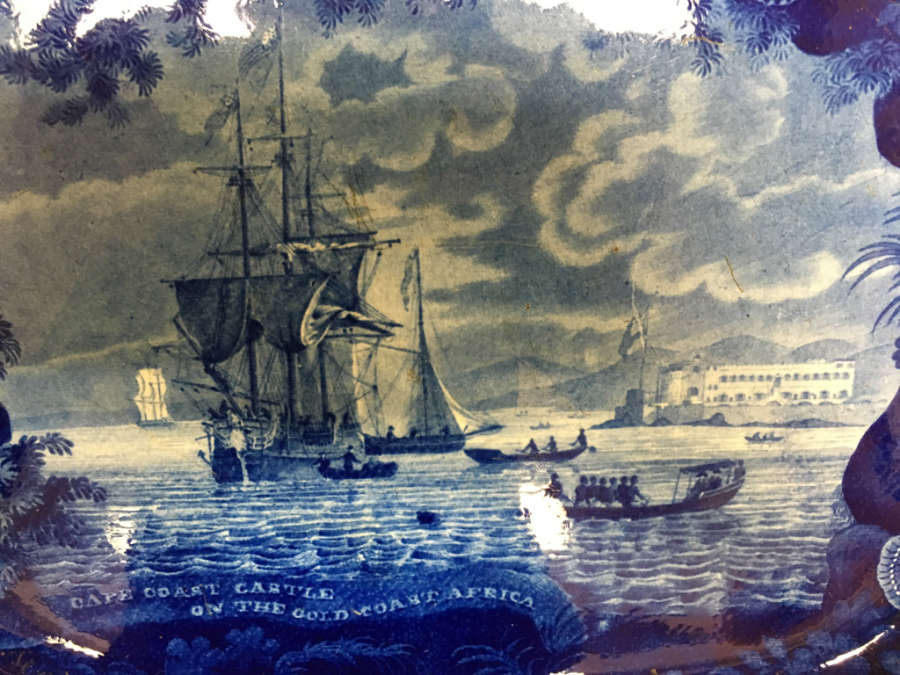 A close up of a blue ceramic illustration depicting a large ship on rough waters under a cloudy sky with text,  "Cape Coast Castle on the Gold Coast, Africa"