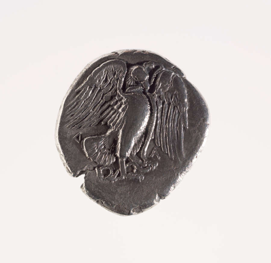 Dull, round, rough-edged chipped silver coin embossed with the image of an upright eagle with spread wings, holding a snake in its claws. Photographed to appear dull.