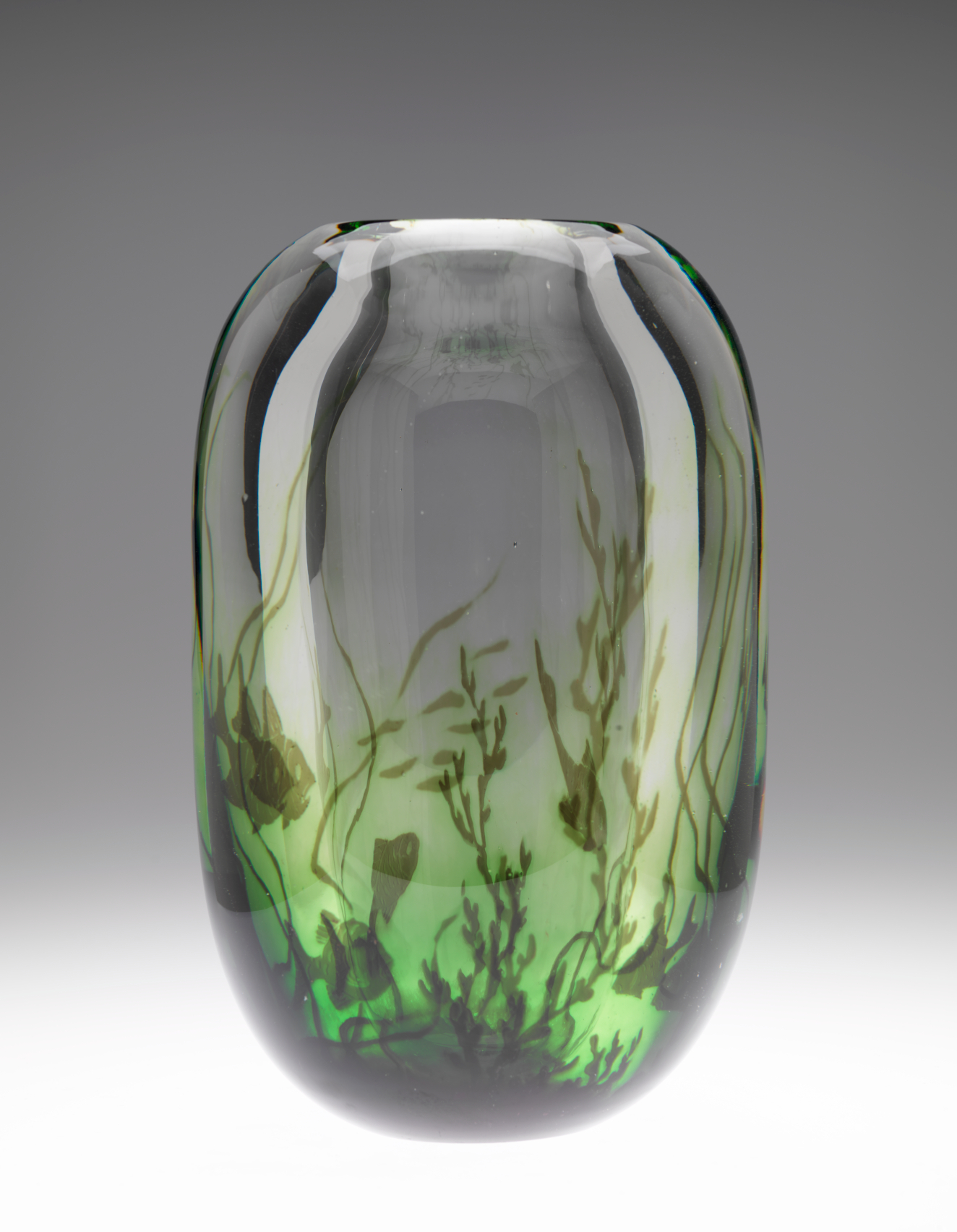 Translucent glass object which transitions to green around the base featuring an underwater scene with fish and aquatic plants. The gray backdrop glows white around the bottom, illuminating the piece.