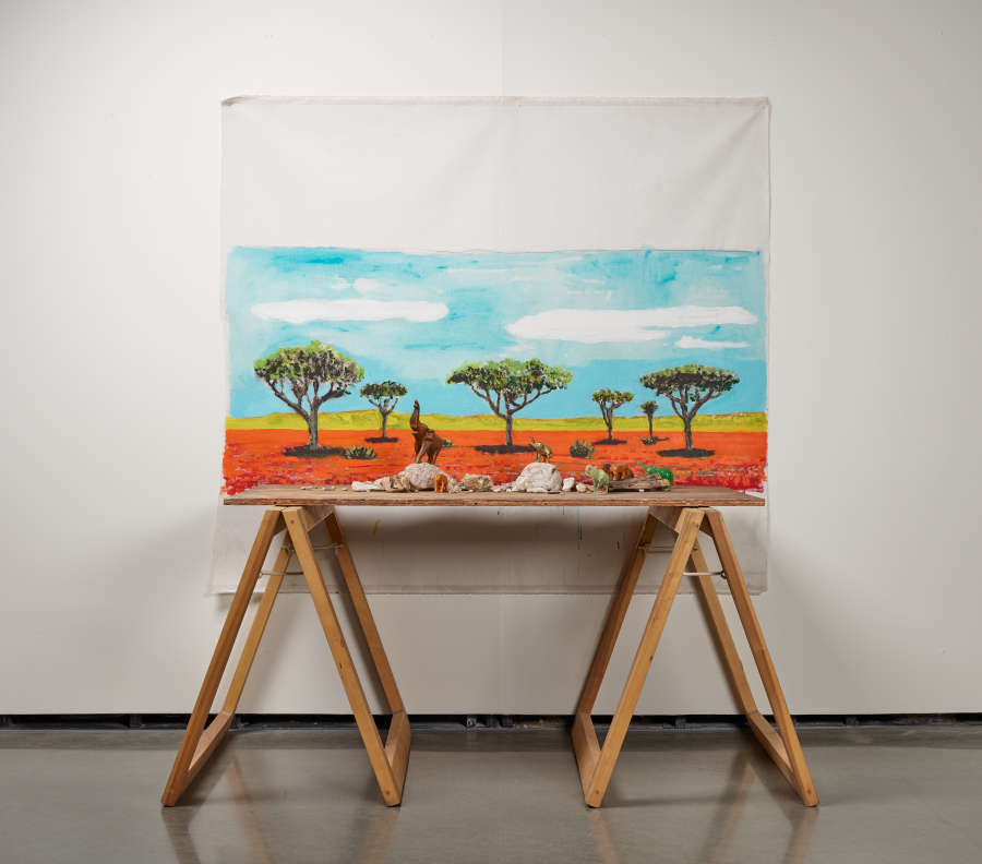 A white cloth painting of six trees in orange soil against a bright blue background with white clouds and green foreground, hung behind a wooden table with various objects.