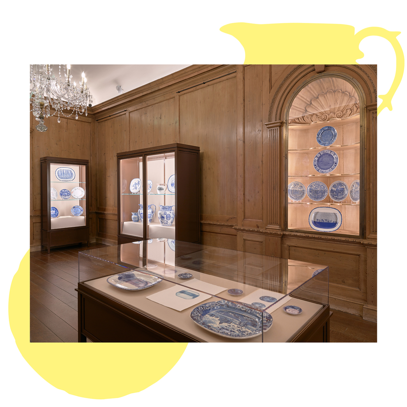 Blue and white ceramics are displayed in cases along the walls of a wood-paneled gallery. A case in the foreground contains additional works. A crystal chandelier hangs at top left.