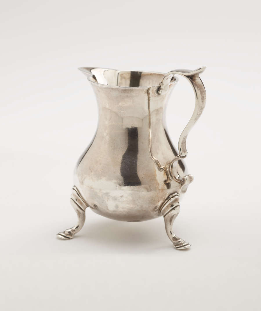 A silver creamer with a sculptural handle and three protruding feet.