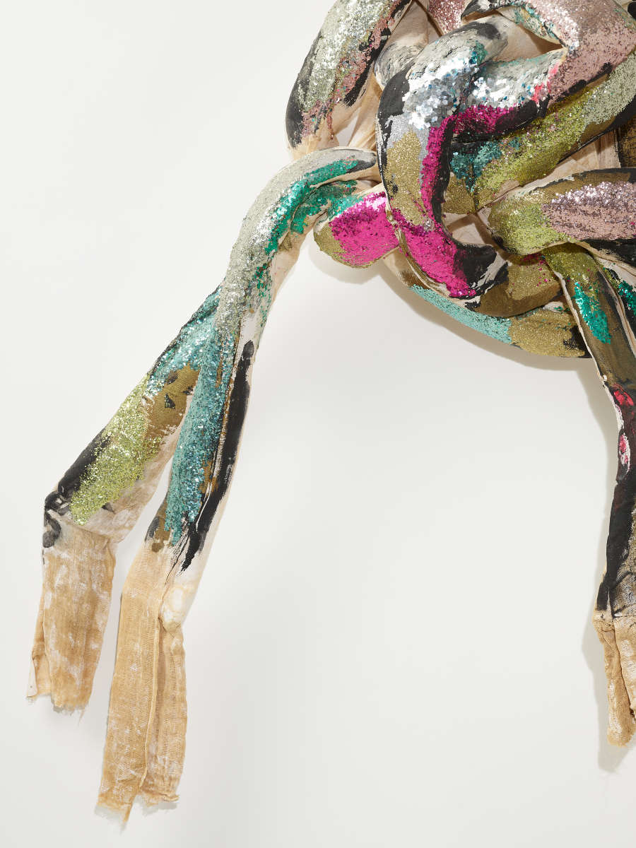 Raw cloth ends of a knotted textile coated in glittering teal, pink, gold, and silver. 