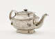 A silver teapot with a decorative handle, a rounded square body and spout with sculptural decorations.