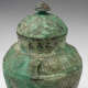 Close-up view of the top of a green jug, with a round bulbous body, short neck, and round lid.  Its green moss-like surface features textured etchings of patterns and characters.