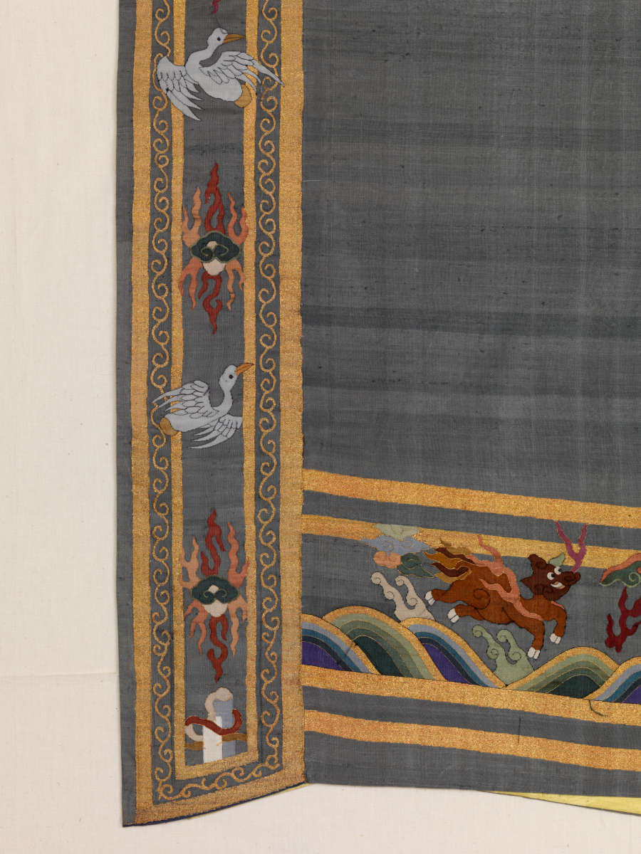 Detail of the blue robe’s sleeve edges featuring animals leaping over waves and vegetal motifs, flying birds and wispy pastel clouds, all encased in perpendicular gold and blue stripe borders.