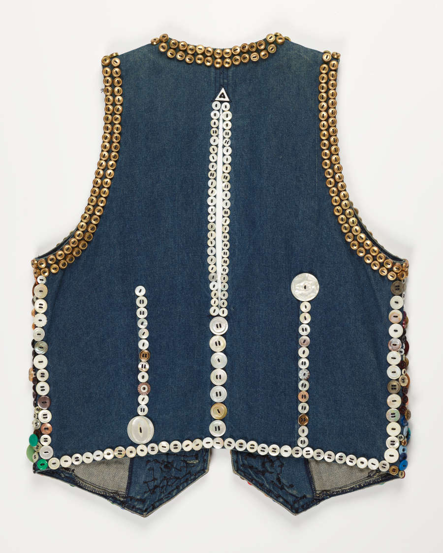 The back of a denim vest decorated with variously sized white and gold buttons. The  ornamentation is arranged in sequential rows down the center and sides, and along the borders.