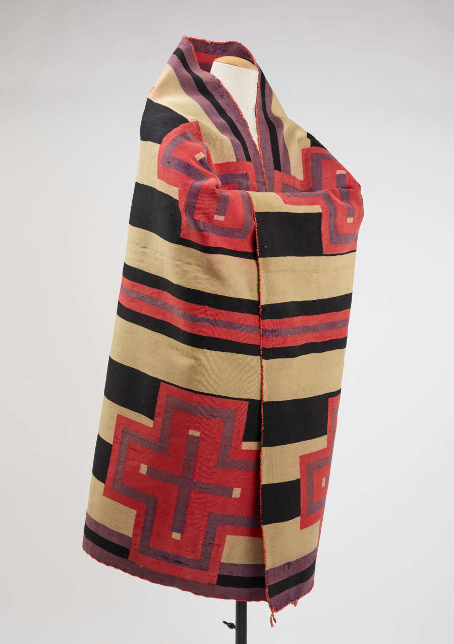 Black and cream striped woven blanket, with pink and purple bordered cross designs, draped around a mannequin. A purple and black stripe lines the collar and bottom of the blanket. 