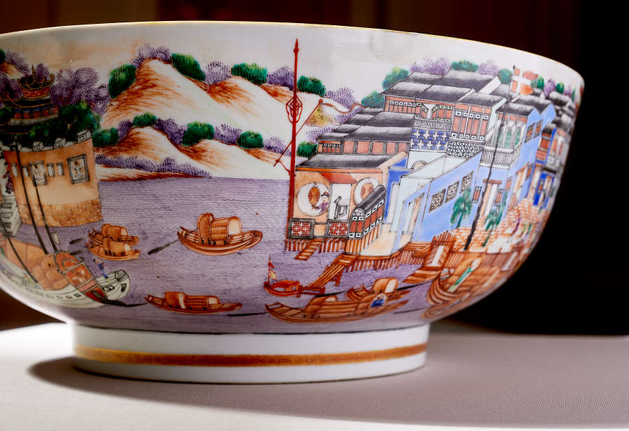 A painted porcelain bowl showcasing intricate designs, including small boats on a purple river, detailed city architecture, and golden hills.