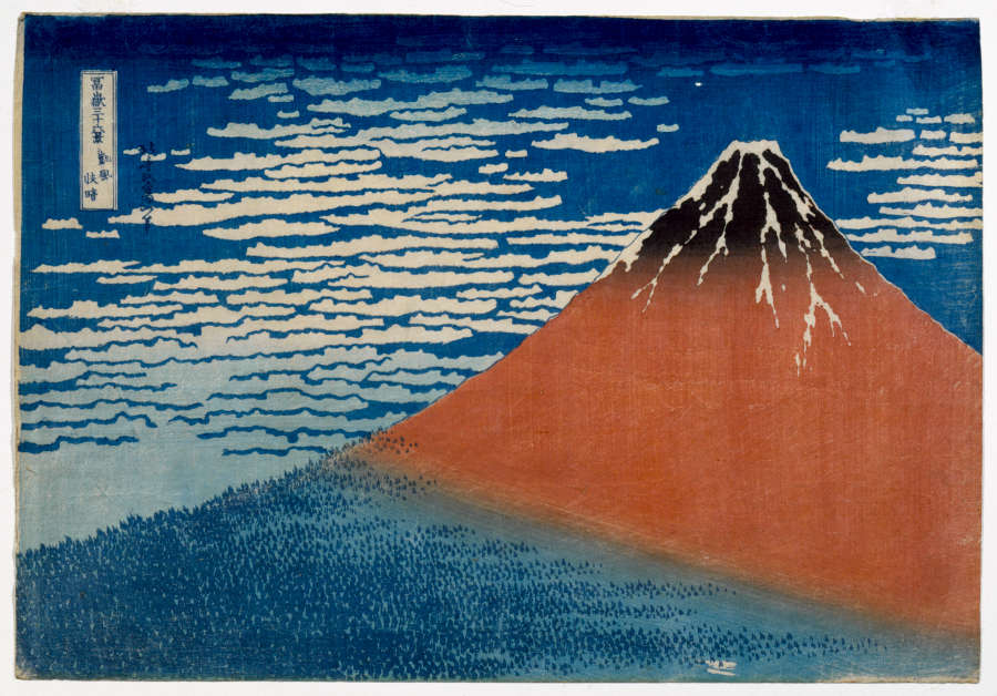 A woodblock print of a partially sunlit mountain against a cloudy blue sky. The mountain's blue shadowed base is forested whilst the red sunlit tip of the mountain is snowy.
