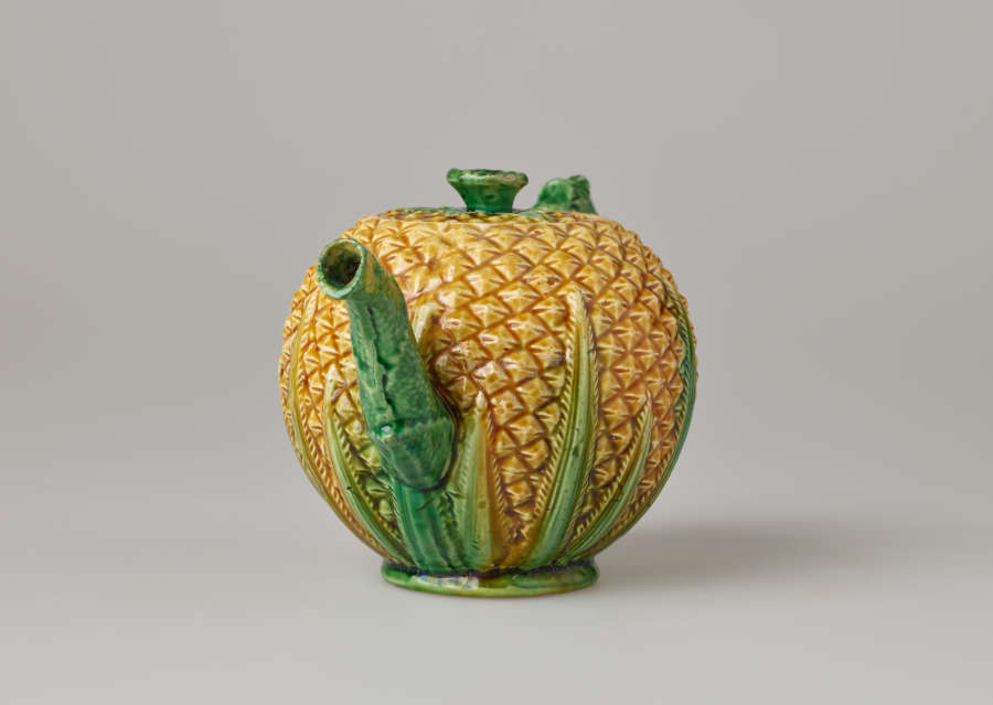 A teapot in the shape of a pineapple. Handle, foot, lid, and spout are green.