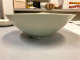 Side view of the glossy gray-green bowl in front of a cluttered counter. Its outside is smooth and uniform with a small base. The top of the plate rises upwards. 