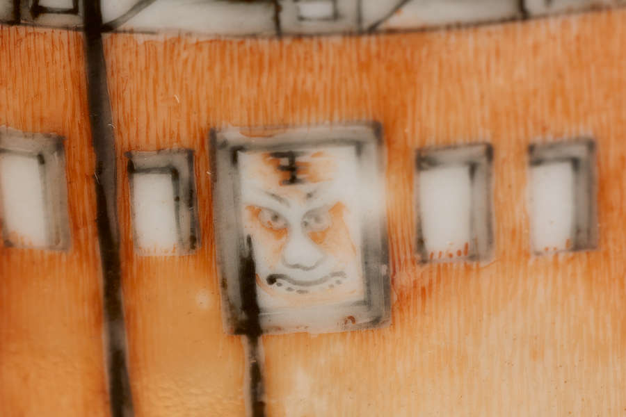 A close up of a painted ceramic depicting a section of an orange wall with small windows, highlighting a rectangular window with an illustrated face and adjacent smaller windows.