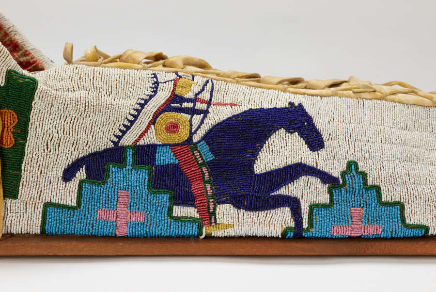 Detail of beaded object. White background beaded with blue horse and colorful rider in headdress, with geometric decoration. Leather lacing at top.