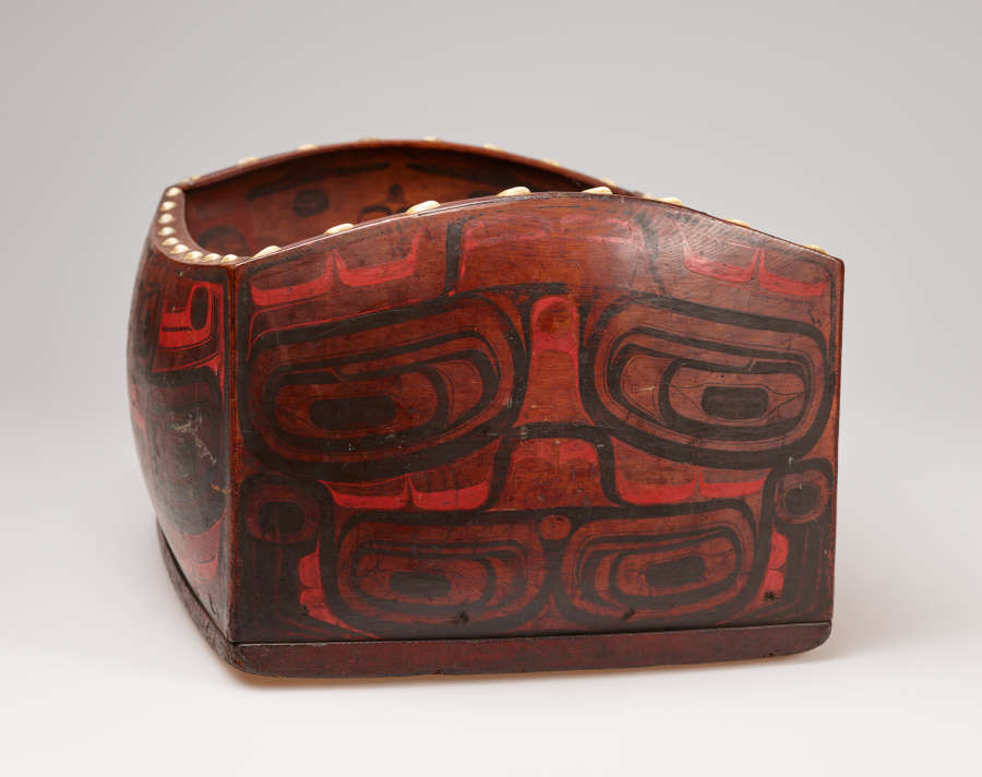 Partial side-view of a rectangular wooden box with bulging sides and faded, red and black, graphic face-like illustrations. The box's rim undulates and has a line of large white dots.