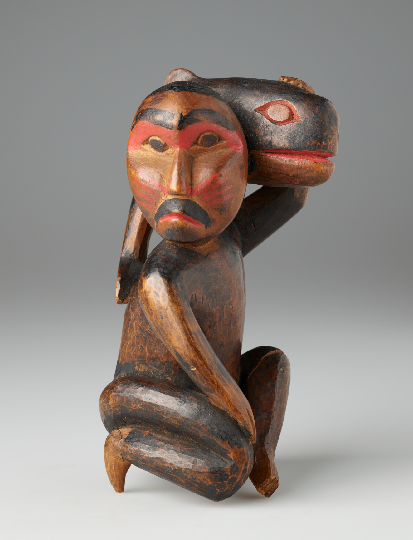 Wooden sculpture of a kneeling bearded man looking aside, holding a small whale against his head. Both man and fish are detailed in partially worn red and black paint.