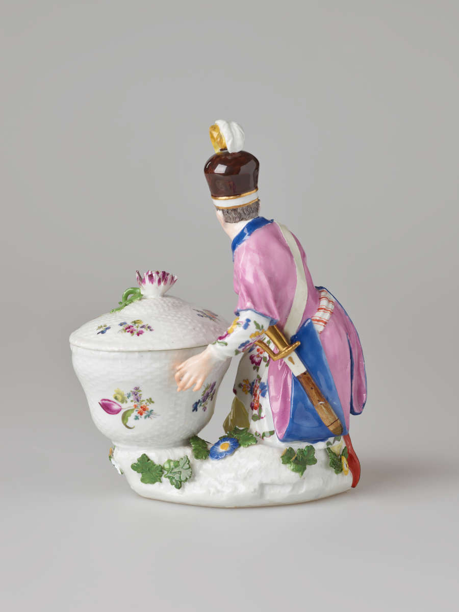 A sculptural figure kneeling next to a basket. Figure wears bright, elaborate clothing with a hat with a feather. There are floral decorations on the figure’s clothing and basket.
