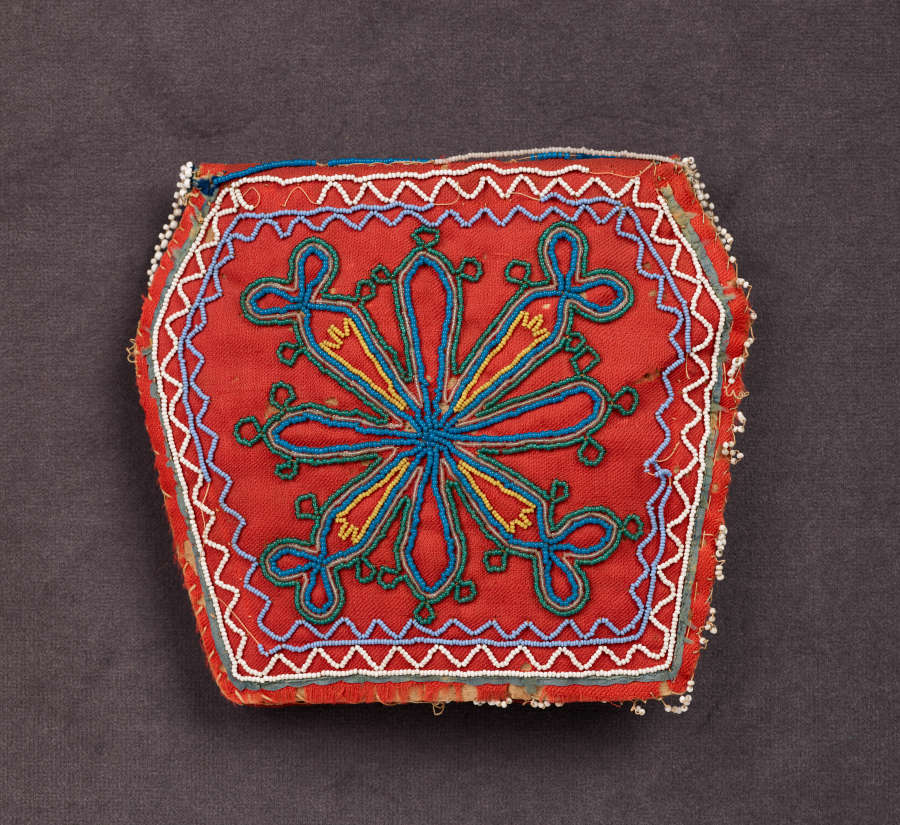 The back of a red irregular hexagonal bag. Wavy triangular patterning in whites and blues are beaded around the border with radial green and blue floral motifs inside.