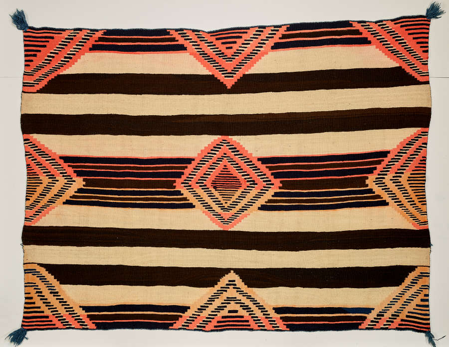 Black and cream striped woven blanket with concentric salmon pink and yellow diamonds overtop. The diamond pattern is cut off along the edges and dark green tassels adorn each corner.