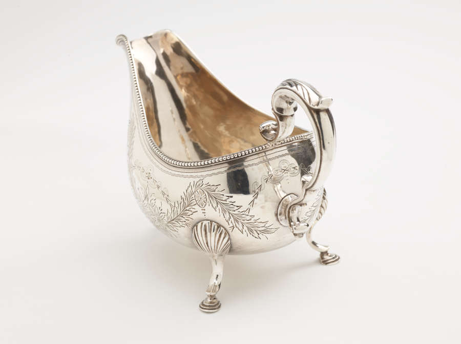 A silver sauce boat with a sculptural handle, high lip for pouring, and three protruding feet. There are floral engravings along the body of the vessel.