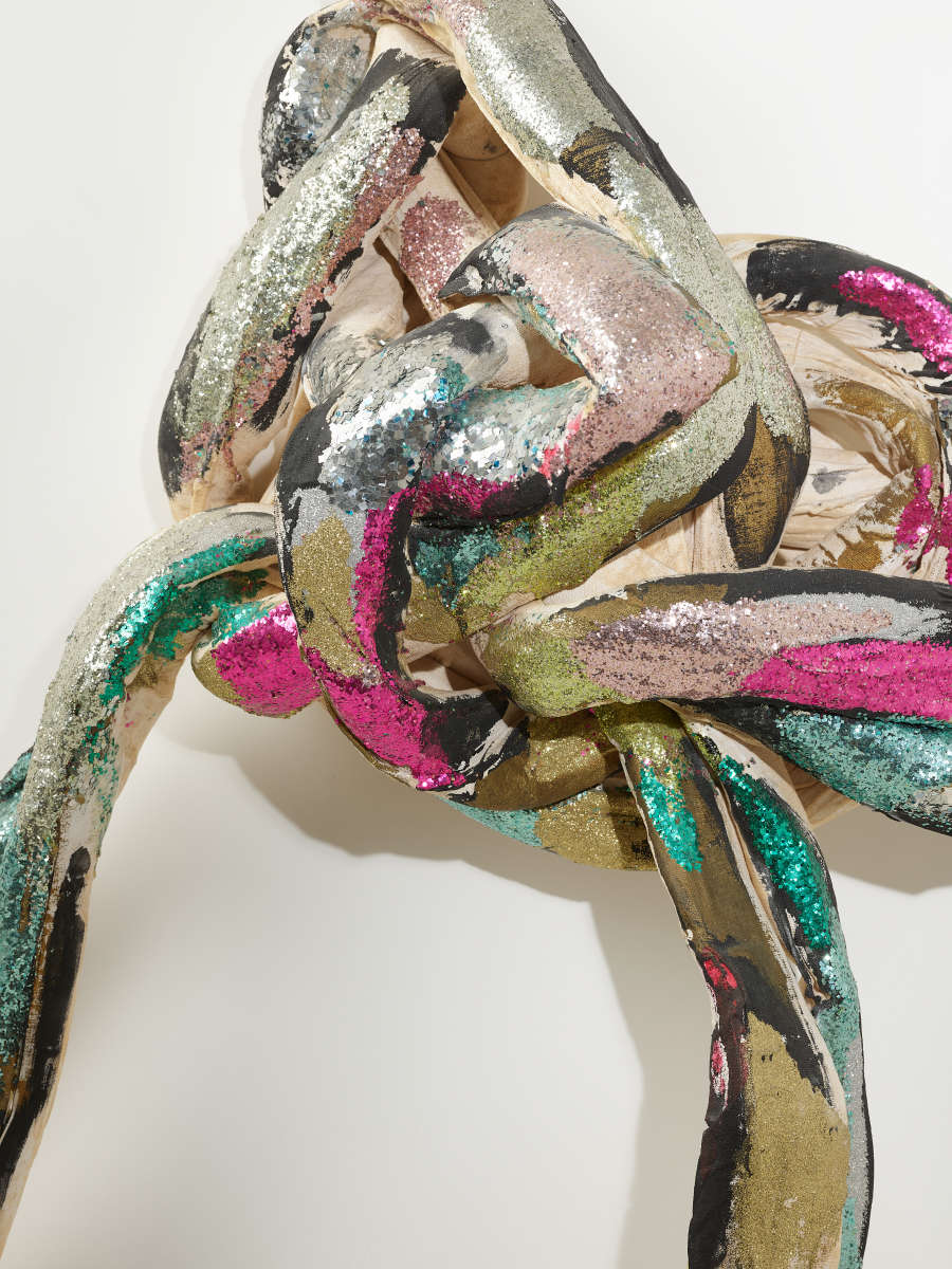 A knotted bundle of textile strips with metallic silver, pink, green, and black cloth, overlapping in a shimmering tangle.