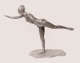 Silver toned sculpture in a bright white environment depicting a female dancer in profile facing to the left balanced on one leg, leaning forward with her other limbs outstretched.