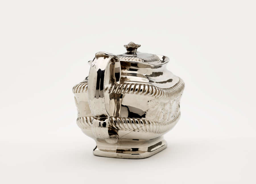 A silver teapot with a decorative handle, a rounded square body and spout with sculptural decorations.