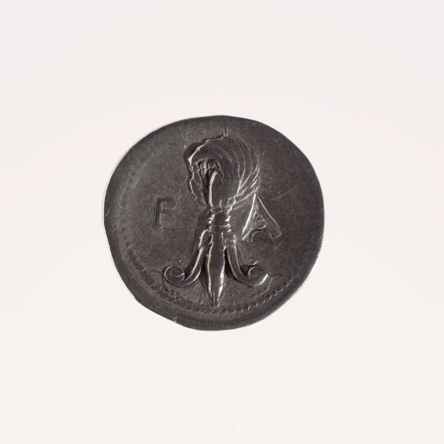 Irregular round silver coin embossed with the image of a feathered fleur-de-lis and the letters “F” and “A” on either side of it. It is framed by a dotted border.